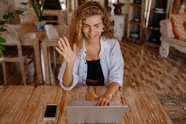 woman seated, smiling and waving at her laptop webcam